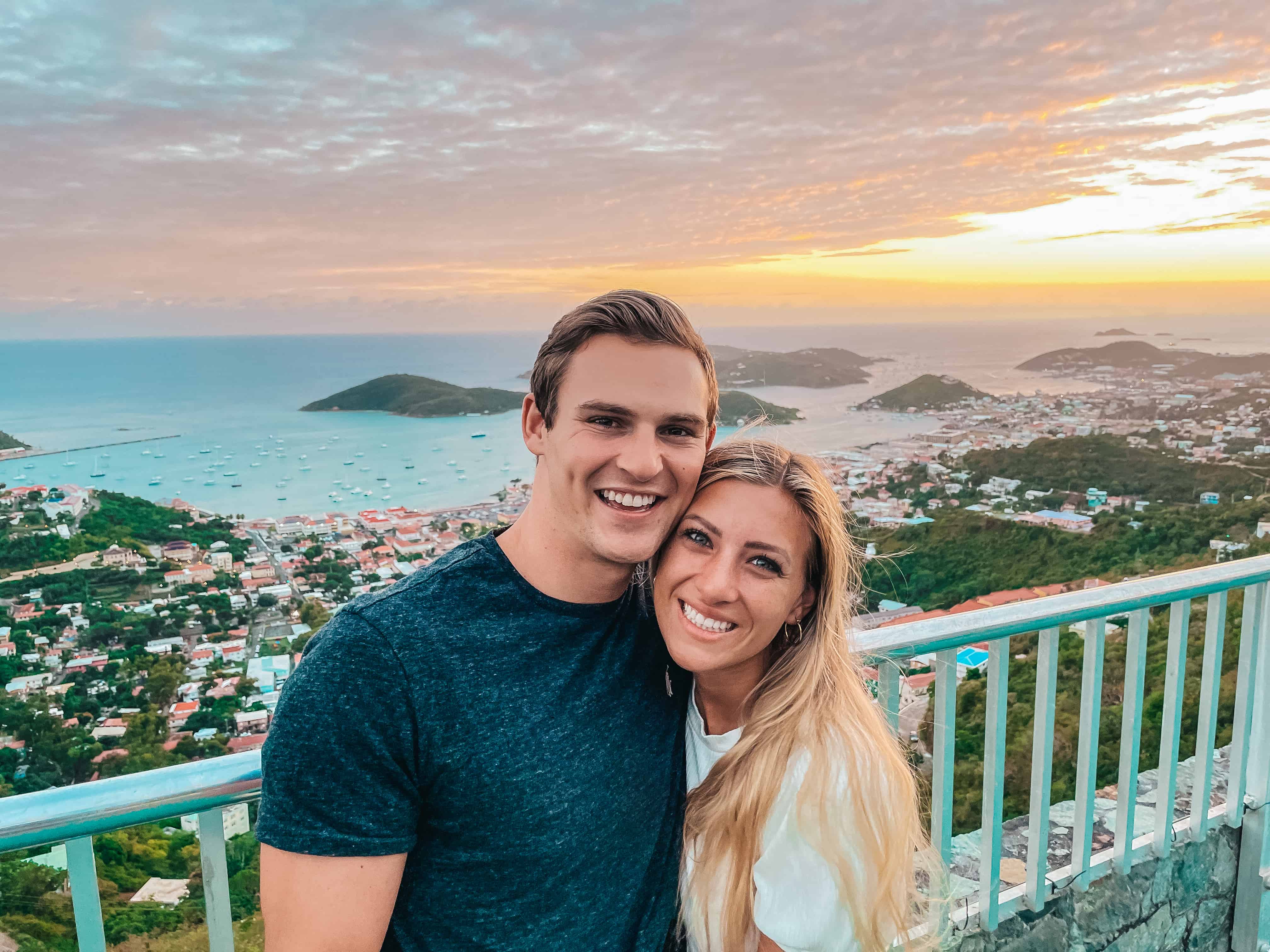 rachel and jason traveling, smiling at the camera at on overlook at sunset over St Thomas with the ocean and green hills